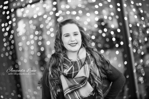 black and white of high school senior girl wearing big scarf standing in front of christmas lights in oklahoma city